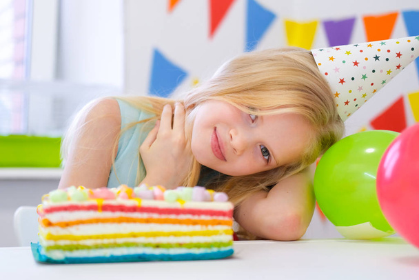 Blonde caucasian girl smiling at camera near birthday rainbow cake. Festive colorful background with balloons - Photo, image