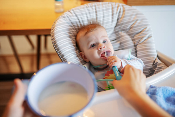 Petit garçon affamé avec un bavoir assis sur sa chaise et une cuillère à lécher. En arrière-plan, sa mère. Quelle délicieuse nourriture
. - Photo, image