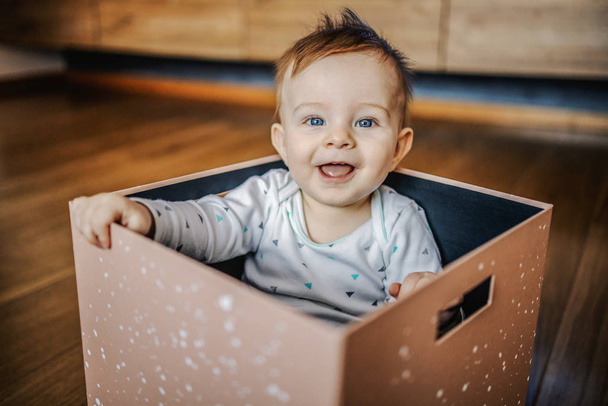 Mavi gözlü sevimli sarışın çocuk kutuda oturuyor, gülüyor, oynuyor ve kameraya bakıyor. İç mimari. - Fotoğraf, Görsel