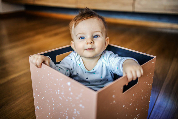 Utelias söpö vaalea valkoihoinen lapsi, jolla on kauniit siniset silmät, istuu laatikossa ja katselee kameraa
. - Valokuva, kuva