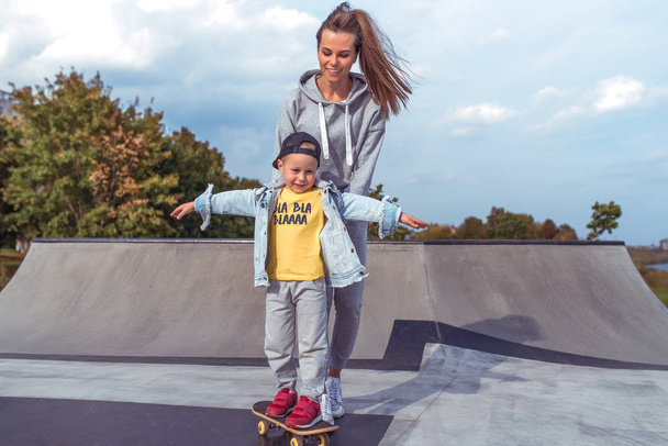 Young family little boy 3-5 years old, woman mother, happy smiling, learning to ride a skateboard, in the summer in a city park. Weekend rest on the sports field. Skateboard casual wear. - 写真・画像