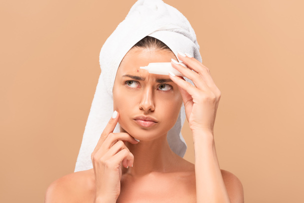 displeased and naked woman in towel holding treatment cream near pimple on face isolated on beige  - 写真・画像