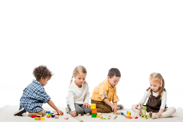wielokulturowe dzieci bawiące się drewnianymi klockami na dywanie, izolowane na białym - Zdjęcie, obraz