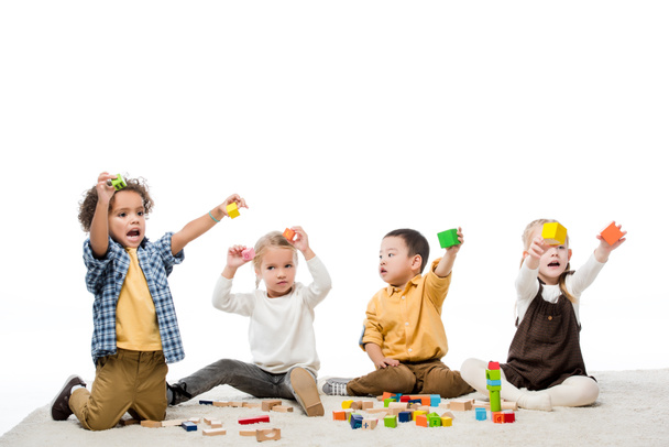 niños emocionales multiculturales jugando con bloques de madera en la alfombra, aislados en blanco
 - Foto, Imagen
