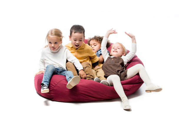 adorable positive multicultural kids playing on red bin bag chair on white - Photo, Image