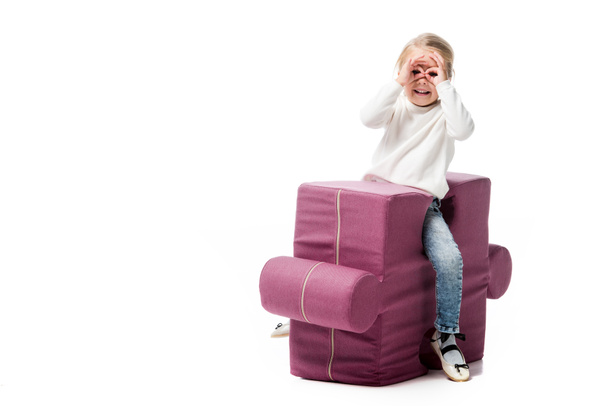 веселый ребенок делает очки из рук на фиолетовом кресле-головоломке, изолированный на белом
  - Фото, изображение