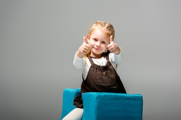 glückliches Kind zeigt Daumen hoch auf blauem Puzzle-Stuhl, isoliert auf grau  - Foto, Bild