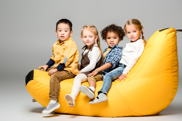 adorable multicultural kids sitting on bin bag chair on grey - Zdjęcie, obraz