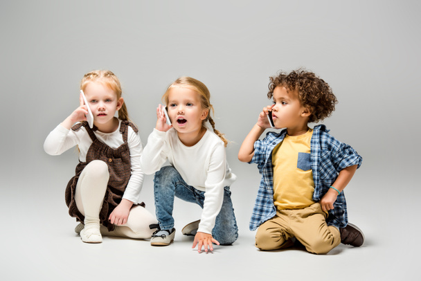 adorable multicultural kids talking smartphones on grey - Foto, afbeelding