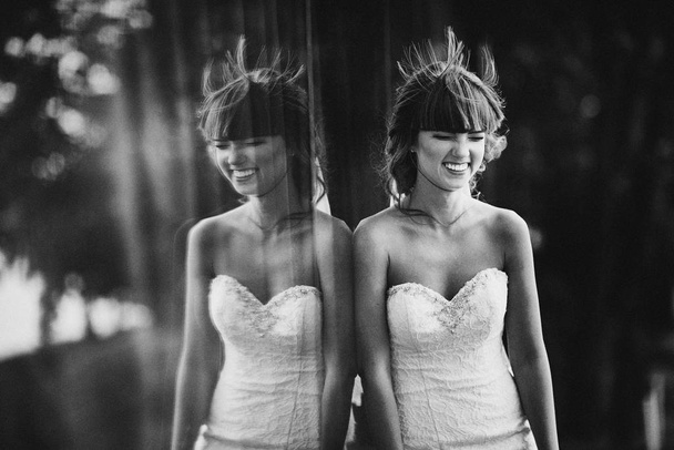 Чорно-біле фото з шумом красивої веселої молодої нареченої в розкішній білій сукні. відображення нареченої у вікні на відкритому повітрі
 - Фото, зображення