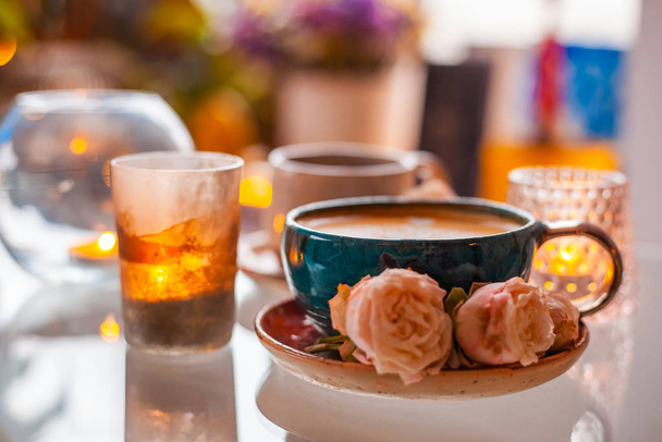 Kuppi kahvia lautasella on koristeltu ruusuilla kynttilöiden vieressä kauniissa kynttilänjaloissa lasipöydällä.
 - Valokuva, kuva