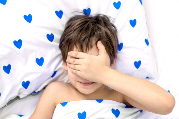 el niño yace en una cama con corazones azules, su cara en sus manos. emociones sin rostro. color blanco, vista superior
 - Foto, Imagen