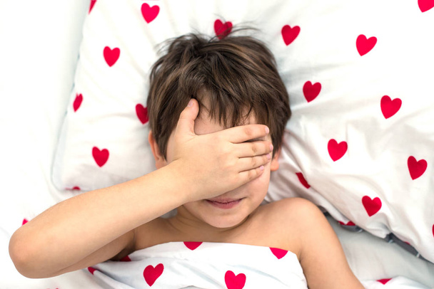 Çocuk kırmızı kalplerle bir yatakta yatıyor, yüzü ellerinde. Yüzü olmayan duygular. beyaz renk, üst görünüm - Fotoğraf, Görsel