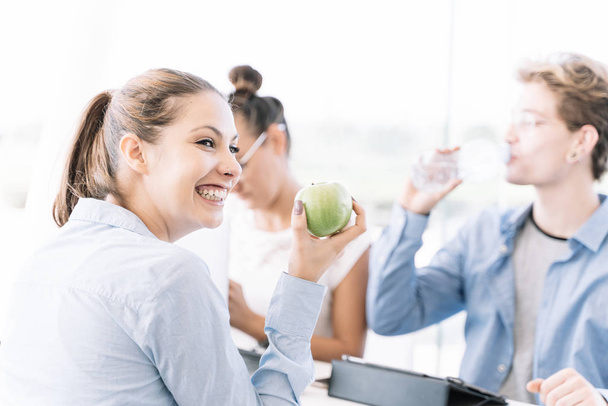 Trois personnes travaillant à une table avec des ordinateurs portables et des tablettes tandis que l'une des filles tient une pomme verte alors qu'elle sourit dans un coworking - Photo, image