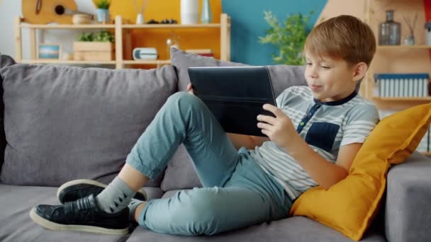 Cheerful little boy holding tablet using modern device on couch at home alone - Кадри, відео