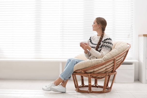 Young woman with hot drink in papasan chair near window at home - Foto, Bild