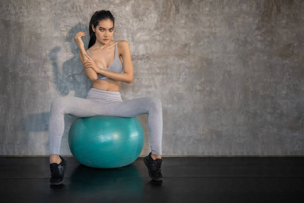 Asiática modelo chica está sentado en una bola de yoga, con un telón de fondo de paredes de hormigón, en el concepto de ejercicio
. - Foto, imagen