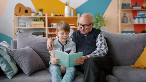 Old man grandfather reading book to smart grandchild indoors in apartment - Filmati, video
