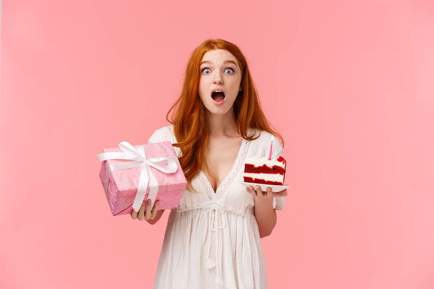 Surprised and curious amazed cute redhead woman reacting to awesome present, seeing incredible thing, holding wrapped gift and peace birthday cake with lit candle, standing pink background - Zdjęcie, obraz