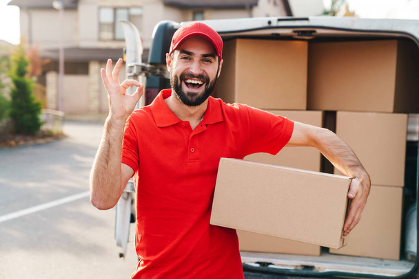Image of a delivery man standing with parcel box and showing ok sign - Foto, afbeelding