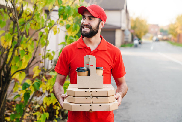 Image of smiling young delivery man holding pizza boxes and coffee - Fotó, kép