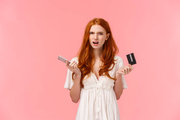 Frustrated and annoyed redhead girl feel displeased with strange online banking system, cant get clue what happened her order, holding smartphone and credit card squinting bothered - Fotografie, Obrázek