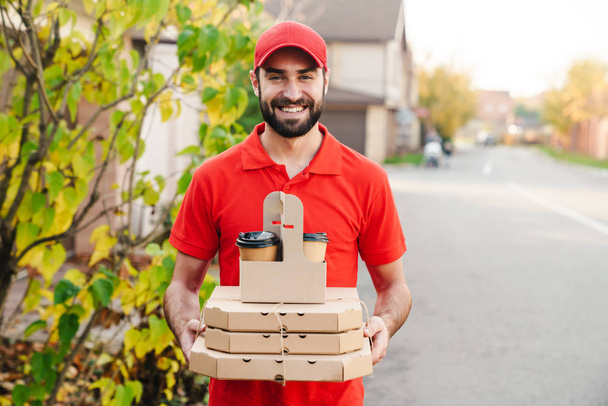 Image of smiling young delivery man holding pizza boxes and coffee - Фото, изображение