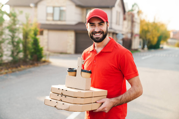 Image of joyful young delivery man holding pizza boxes and coffee - Fotografie, Obrázek