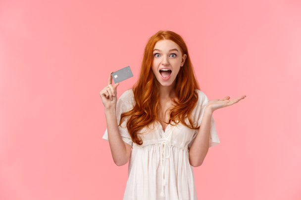 Surprised and impressed cute happy, cheerful redhead girl amazed she got awesome bonuses on bank account, using credit card, saved-up money on deposit or cashback, pink background - Photo, Image