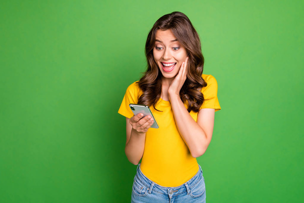 Photo of beautiful wavy lady holding telephone hands reading website blog new positive comments wear casual yellow t-shirt jeans isolated bright green color background - Fotó, kép