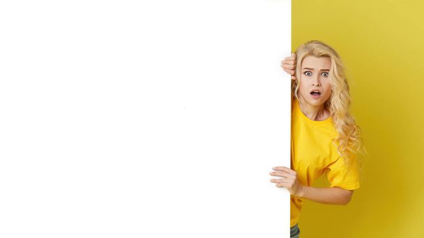 Young happy woman peeks out from behind a white banner on a yellow background. Point to an empty blank on a form, a copy space for text. Horizontal shot - Foto, Imagem