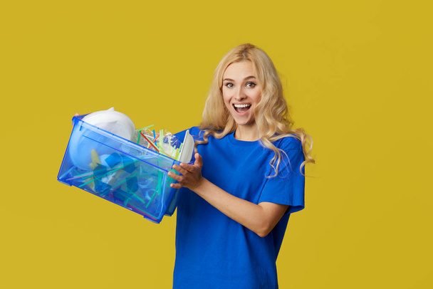 Caring for nature. Young woman in a blue T-shirt are holding a box with plastic bottles and waste. Volunteer compiled garbage and carry it to recycling. Yellow background - Foto, afbeelding