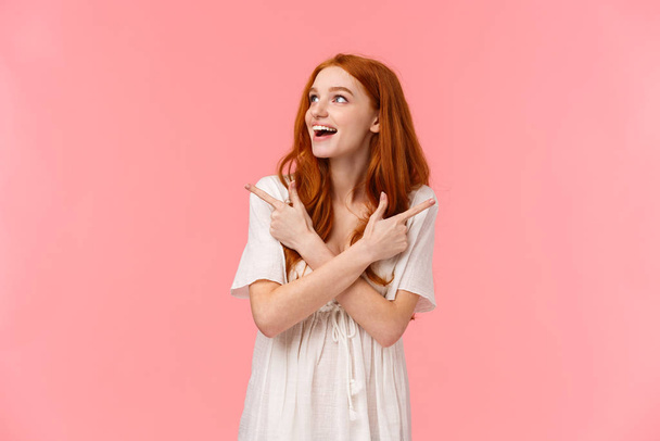 Dreamy attractive redhead female, looking around wondered and amused with happy smile, glance left as pointing sideways, left and right products, making final choice what buy, pink background - Photo, image