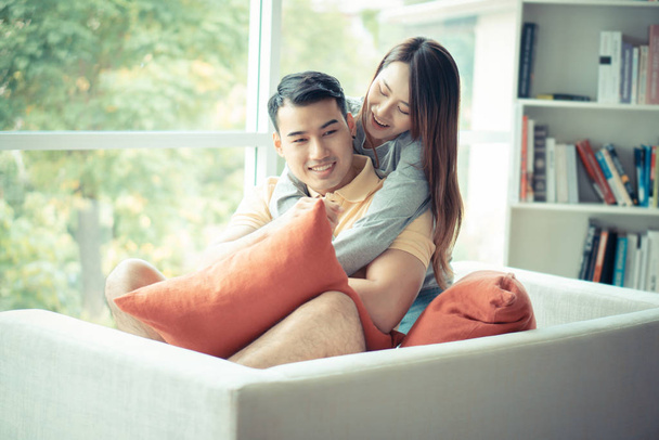 glückliches Paar, das auf dem Sofa sitzt und eine Frau ist, die ihren Freund im Wohnzimmer liebevoll umarmt und lächelt. Konzept der Romantik am Valentinstag. Heiratsantrag und Ehe - Foto, Bild