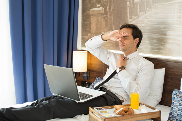 Müder Geschäftsmann im Anzug arbeitet am Hotelbett auf seinem Computer - Foto, Bild