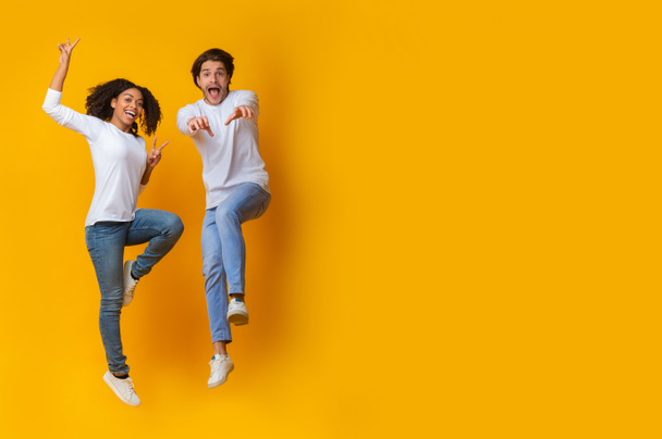 面白い異人種間のカップルは、スタジオで黄色の背景に空気中でジャンプ - 写真・画像