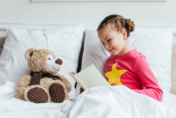 adorable child reading book to teddy bear while sitting on bed - Фото, зображення