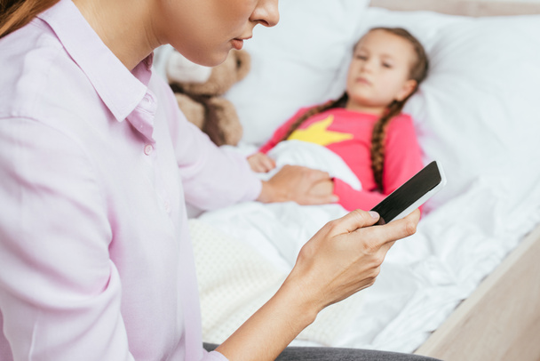 bijgesneden weergave van moeder met smartphone met zieke dochter in bed  - Foto, afbeelding