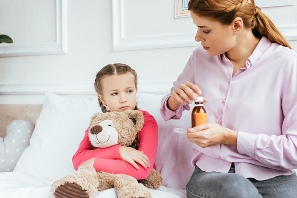 mother giving syrup to sad ill daughter with teddy bear - 写真・画像