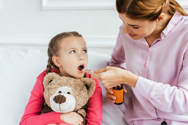 mother giving syrup to ill daughter with teddy bear - Photo, Image