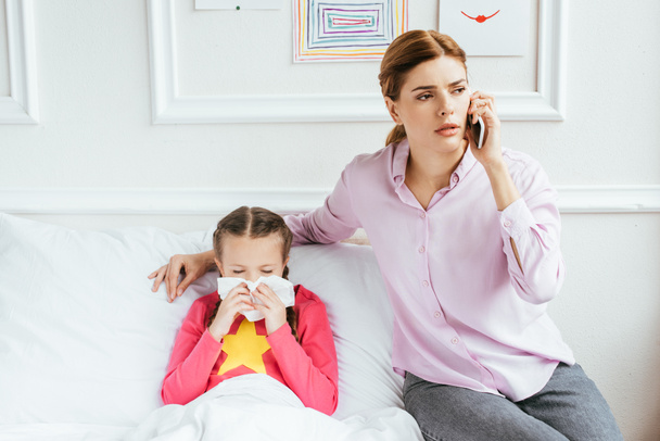 Besorgte Mutter mit Online-Beratung auf Smartphone, während kranke Tochter mit laufender Nase im Bett sitzt - Foto, Bild