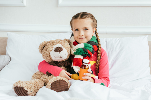 niño enfermo sonriente en bufanda sosteniendo jarabe y sentado en la cama con oso de peluche
 - Foto, Imagen