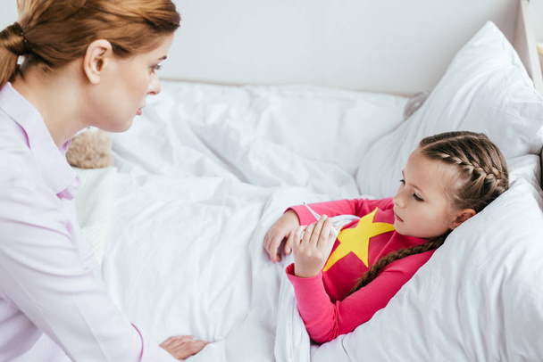 Kranke Tochter hat Fieber und schaut auf Thermometer, während sie mit Mutter in der Nähe im Bett liegt  - Foto, Bild