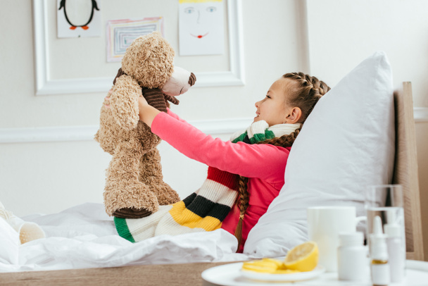 счастливый больной ребенок в шарфе держа плюшевого мишку на кровати с лекарствами рядом
 - Фото, изображение