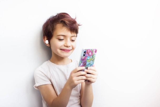 Retrato horizontal pouco 7 anos de idade estudante de cabelos curtos com cabelo rosa ouvindo música e olhando no telefone celular com fones de ouvido sem fio brancos. Estilo de vida inteligente na infância. Viciado em gadgets
 - Foto, Imagem