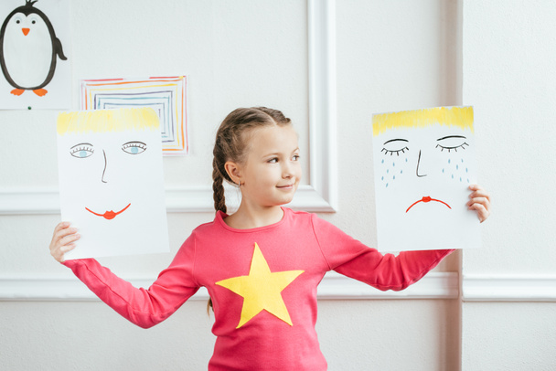 smiling child holding two paintings with happy and sad faces - Φωτογραφία, εικόνα