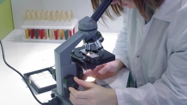 Young girl scientist looking through a microscope. She studies the samples. A young scientist conducts scientific research. She is researching a virus, looking for a cure for an epidemic vaccine - Imágenes, Vídeo