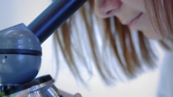 Young girl scientist looking through a microscope. She studies the samples. A young scientist conducts scientific research. She is researching a virus, looking for a cure for an epidemic vaccine - Filmati, video