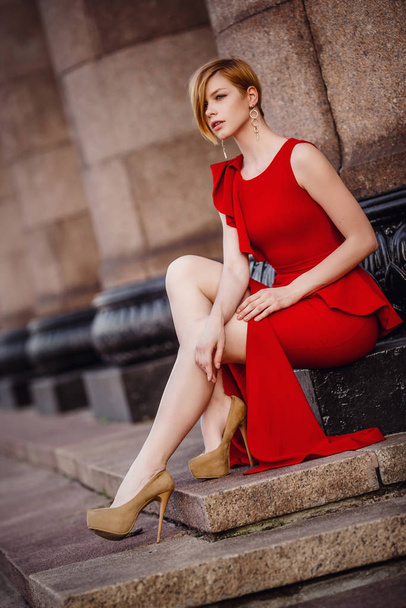 Uzun kırmızı elbiseli, yüksek topuklu ayakkabılı zarif bir kadın merdivenlerde oturuyor. - Fotoğraf, Görsel