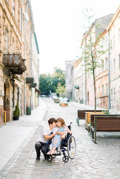 Couple in love in old city center. Pretty young girl with disease on a wheelchair and her lovely man, walking, smiling and touching foreheads, closed eyes - Zdjęcie, obraz